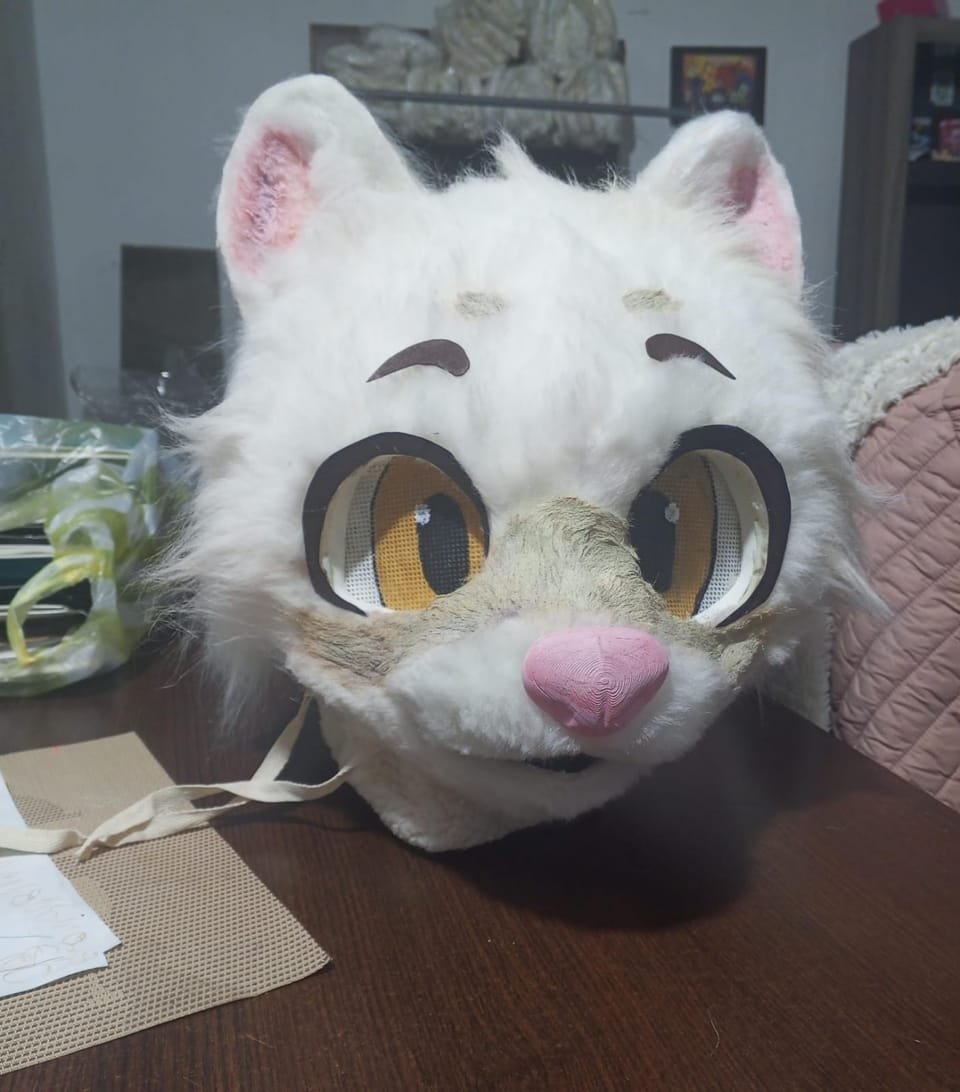 Cabeza de fursuit de gato blanco con ojos naranjas y nariz rosa, sobre mesa de madera.