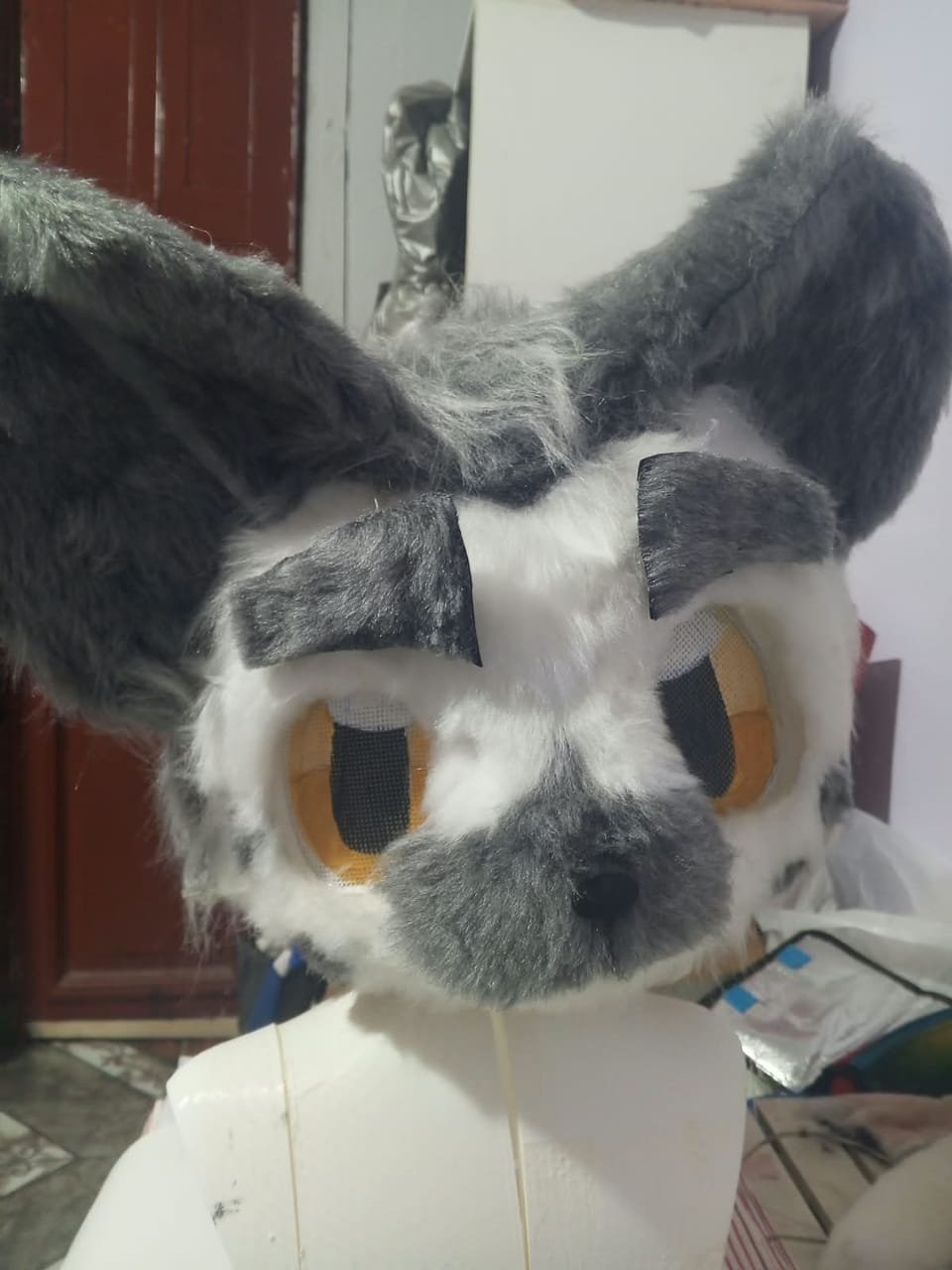 Cabeza de fursuit gris y blanca con orejas extendidas y ojos naranjas, sobre maniquí.