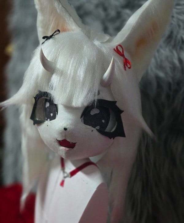 Cabeza de fursuit blanca tipo zorro espiritual, primer plano del rostro con ojos de malla y moño rojo.